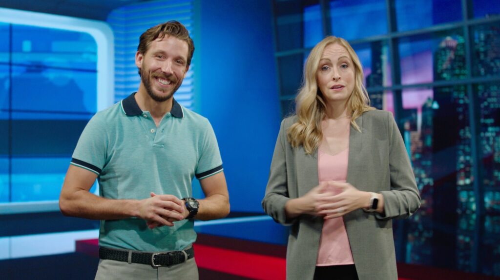 Two presenters standing in a brightly lit studio, one smiling confidently and the other speaking, with a modern backdrop featuring cityscapes and screens.