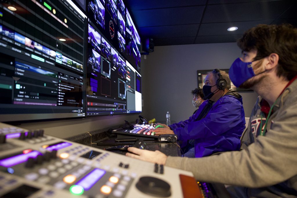 A production team in a control room operating broadcast equipment and monitoring multiple screens, with staff wearing masks for safety precautions.