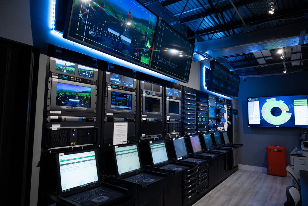 A high-tech server room with multiple monitors, laptops, and racks displaying live broadcasts and data, illuminated by blue LED lighting for a modern production setup.