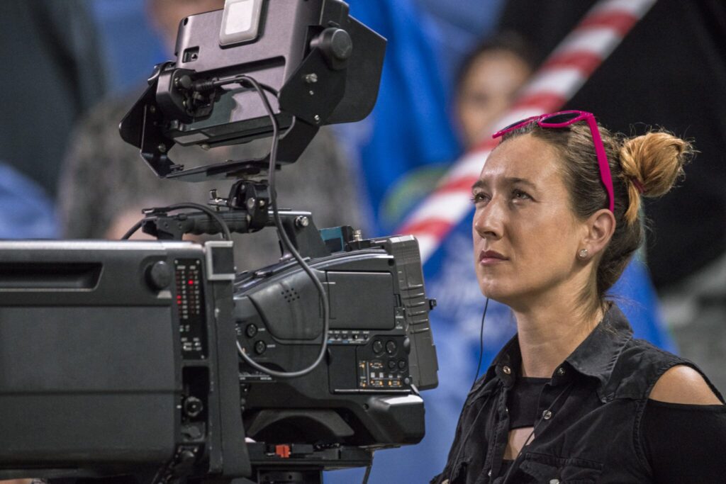 A focused camera operator wearing pink sunglasses on her head, standing behind a professional broadcast camera during an event.