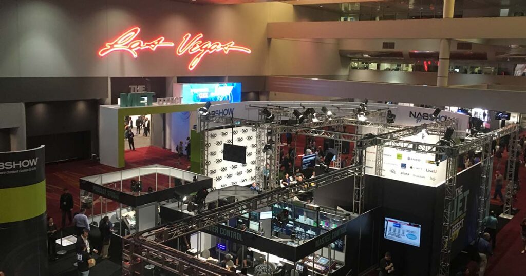 A bustling trade show floor at the NAB Show in Las Vegas, featuring exhibition booths, lighting rigs, and attendees networking under a glowing "Las Vegas" neon sign.