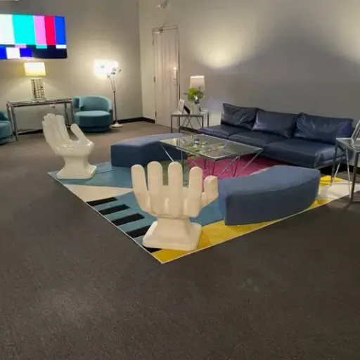 A stylish lounge area with modern furniture, including a navy-blue leather sofa, two unique hand-shaped chairs, and a colorful geometric rug. The space is illuminated by floor lamps and table lamps, with a television displaying a test screen mounted on the wall.