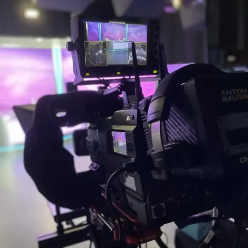 A professional video camera on a set, equipped with a monitor displaying a live view of a brightly lit studio featuring a purple backdrop. The camera setup includes advanced recording equipment, indicating a high-end production environment.