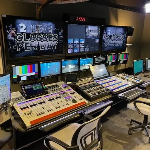 A fully equipped broadcast control room featuring multiple workstations with chairs, large monitors displaying live production feeds, colorful test screens, and video editing equipment. The setup is organized and modern, reflecting a professional production environment.