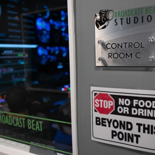 A control room entrance at Broadcast Beat Studios with a wall-mounted sign reading "Control Room C" and a warning below stating "No Food or Drink Beyond This Point." Through the glass, a high-tech control room setup with monitors and a person operating equipment is visible.