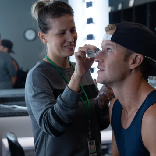 Makeup artist applying makeup to a man wearing a backward cap in a dressing room, with bright vanity lights and a relaxed atmosphere in the background.