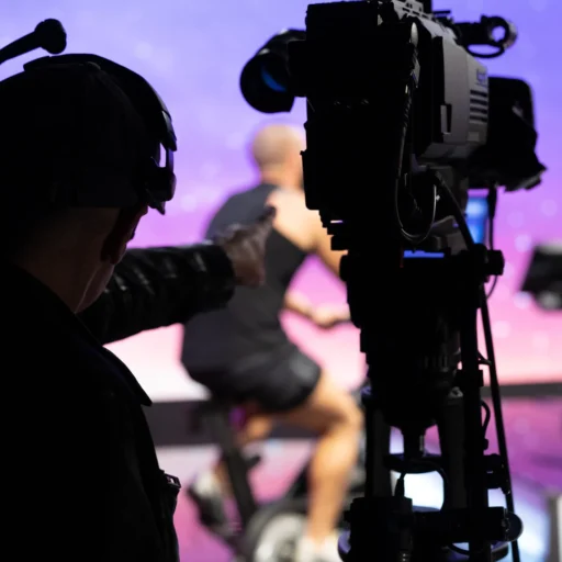 Close-up of a camera operator wearing a headset, pointing towards a live fitness studio set where individuals are exercising against a vibrant purple backdrop. A professional video camera is in focus, capturing the scene.