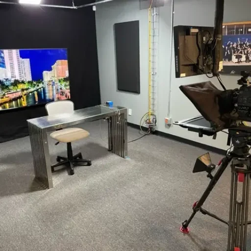 Small broadcast studio setup with a sleek metallic desk and chair, a large monitor displaying a cityscape, and a professional camera on a tripod equipped with a teleprompter.