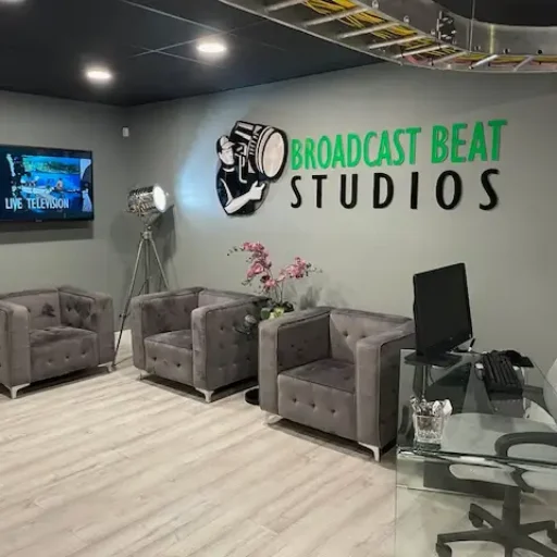 Modern studio reception area featuring gray tufted chairs, a glass desk with a computer, a television on the wall, and the Broadcast Beat Studios logo prominently displayed.